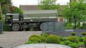 Samochód ciężarowy z napisem patrol saperski stoi przed bramą wjazdową na posesję
