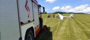 Wóz strażacki na pierwszym planie, przed nim szybowiec na zielonej łące