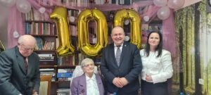 Setne urodziny Pani Anieli Koreckiej – jubilatka siedzi na fotelu, obok niej przedstawiciele samorządu, nad głową złoty napis „100