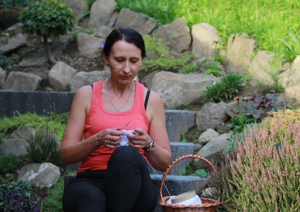 młoda kobieta o czarnych włosach i różowej koszulce bez ramion siedzi na kamiennych schodach i robi na szydełku