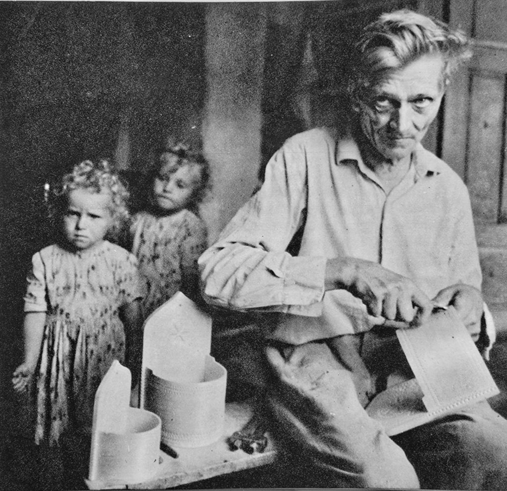 Czarno-białe zdjęcie starszego mężczyzny rzeźbiącego w drewnie. Za nim stoją dwie kilkuletnie dziewczynki