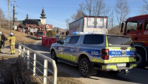 Samochody służb ratunkowych i policyjnych przy miejscu wypadku w Rdzawce, ustawiony parawan strażacki zasłania miejsce zdarzenia, a w tle na horyzoncie widoczny jest kościół.