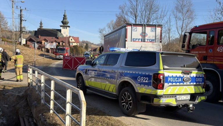 Samochody służb ratunkowych i policyjnych przy miejscu wypadku w Rdzawce, ustawiony parawan strażacki zasłania miejsce zdarzenia, a w tle na horyzoncie widoczny jest kościół.