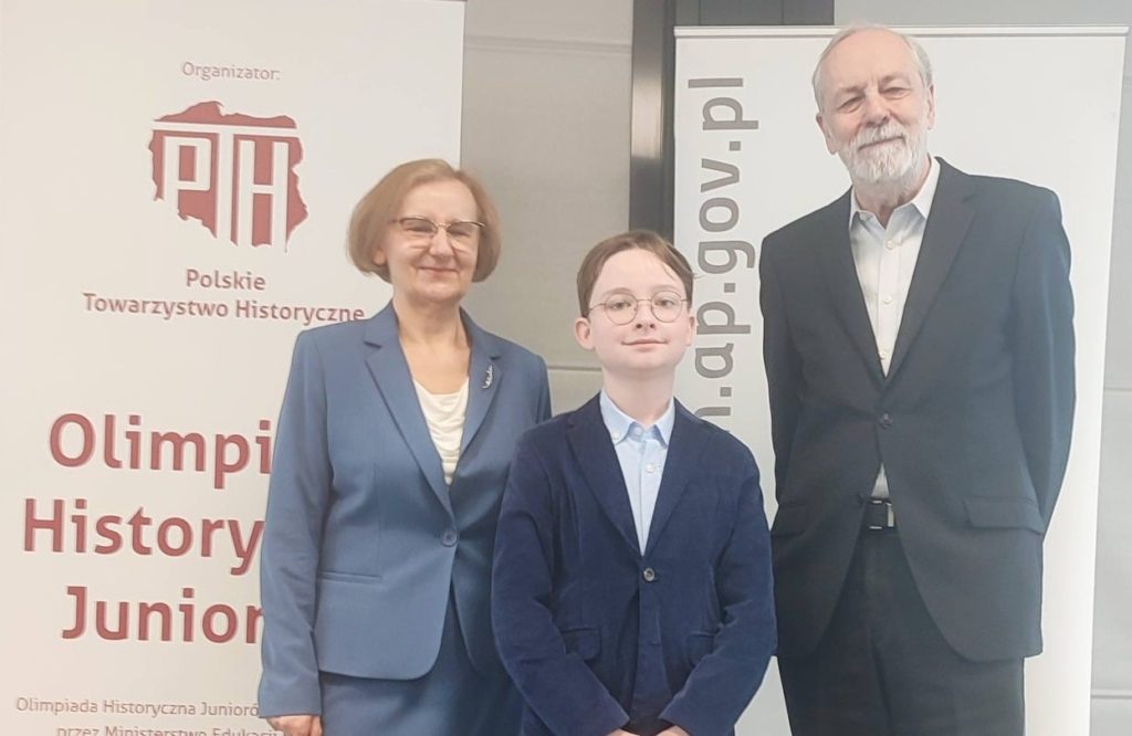 Wojciech Miśkowicz stoi w towarzystwie dwóch dorosłych osób na tle plakatu